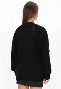 Juodas megztinis su laisvu kirpimu, su pašoniniu ir rankovių raštu. Kiekvieną rankovę puošia dekoratyvinis raudonas pom-pom. Audinys su tekstūru.