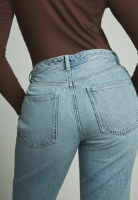 Jean en denim bleu clair avec une taille haute, des poches arrière et un léger dégradé. La texture est lisse avec des coutures piquées et un léger effet usé.