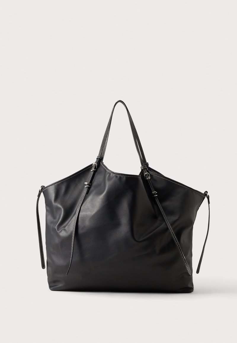 Grand sac fourre-tout en cuir noir avec doubles bretelles et liens latéraux réglables, sur un fond clair uni.