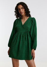 Groene wikkeljurk met puffy mouwen, textuurstof en een strikdetail in de taille. Het patroon heeft een ruitontwerp. Korte lengte.