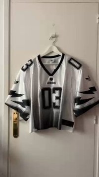 Balta futbola krekls ar melniem akcentiem, kurā ir numurs "03" un adidas zīmols. Īsas piedurknes, sietveida tekstūra, pakārts uz āķa.