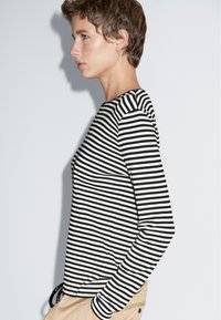 Schwarz-weißer gestreifter Langarmoberteil mit rundem Halsausschnitt, entspannter Schnitt und dezenten Nahtdetails. Kombiniert mit beigen Hosen.