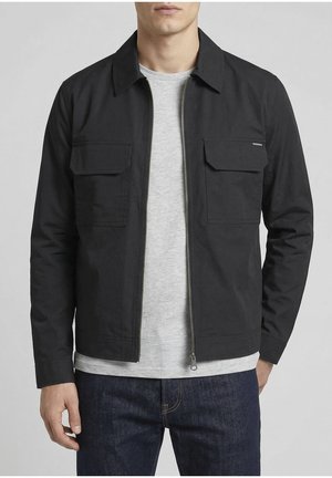 Hombre con una chaqueta negra con cremallera y bolsillos con solapa en el pecho, sobre una camiseta gris claro y jeans azul oscuro.