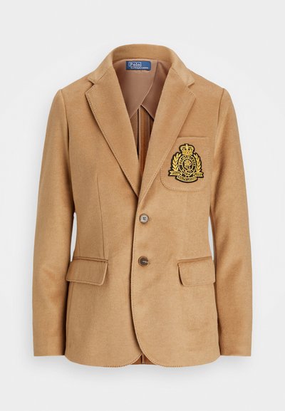 Dzeltenīgs bleizeris ar iegrieztu apkakli, divām pogām un krūšu kabatu. Tam ir izšūta logo zeltā. Audums šķiet mīksts un teksturēts.