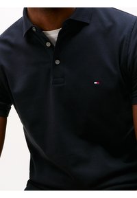 Navy polo-paita, joka on valmistettu teksturoidusta kankaasta, jossa on kolmen napin läpivienti ja pieni logo punaisella, valkoisella ja sinisellä rinnassa.