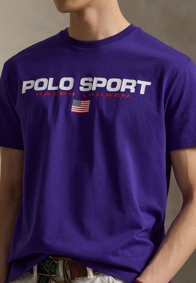 T-shirt en coton violet avec un grand texte « POLO SPORT » en blanc et rouge, un petit drapeau américain en détail, et un col rond.