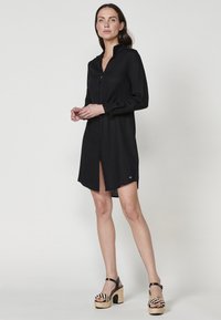 Rochie neagră, cu nasturi, cu mâneci lungi, croială dreaptă și slituri laterale. Purată cu sandale cu platformă negre și bej, având un design împletit.