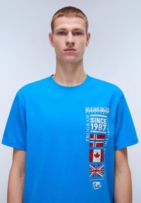 Modré tričko s kulatým výstřihem vyrobené z bavlny s grafickým potiskem, na kterém jsou vlajky a text: "NAPAPIJRI OD ROKU 1987" a "PROGRAM ANTARKTIDA."