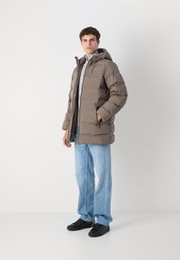Bruine gewatteerde jas met capuchon, voorzien van gestikte delen, dubbele zijzakken en een grijze voering, gedragen over lichtblauwe jeans en zwarte sneakers.