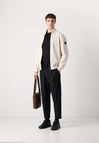 Giovane uomo con una giacca bomber crema, camicia nera, pantaloni neri e sneakers, che tiene una borsa a tracolla marrone davanti a uno sfondo bianco.