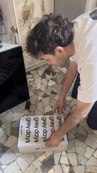 White rectangular box with black text "grey days," being lifted on a patterned tiled floor. Hands are securing the box.
