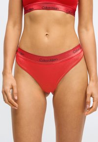 Sarkans kokvilnas maisījuma bikinis ar gludu virsmu, kas aprīkots ar plašu melnu zīmolu jostu ar "Calvin Klein" logo gar malu.