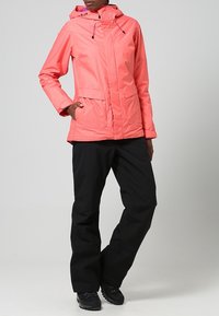 Korallfärgad vattentät jacka med huva, som har två framfickor och en subtil randig textur, kombinerad med svarta byxor och stövlar.