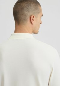 Polo shirt crema in tessuto texture, con colletto classico e maniche corte. La vista posteriore mostra un design pulito e semplice.