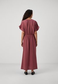 Robe longue et ample en tissu doux et texturé de couleur rose poussiéreux, avec des manches courtes et une taille élastiquée. Des chaussures noires complètent la tenue.