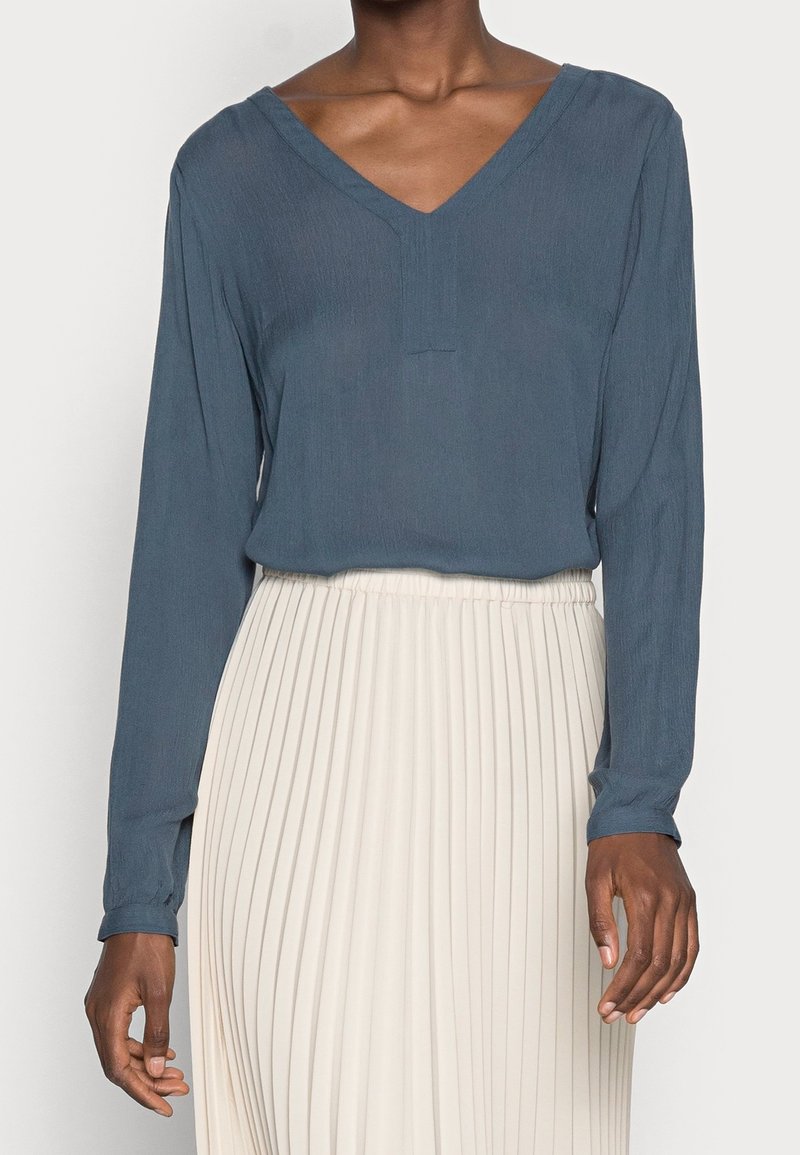 Femme portant un haut bleu à manches longues avec un col en V, rentré dans une jupe blanche plissée taille haute, raccourcie à mi-torse.