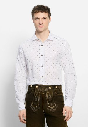 Mann trägt weißes Hemd mit kleinen blauen Mustern und dunkelbraune bestickte traditionelle Lederhose, steht vor weißem Hintergrund.