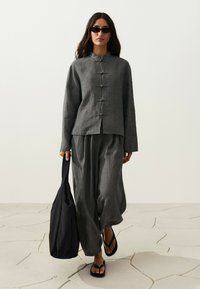 Femme portant une veste grise ample et un pantalon avec des lunettes de soleil noires, portant un grand sac cabas noir et marchant sur un sol en pierre fissuré.
