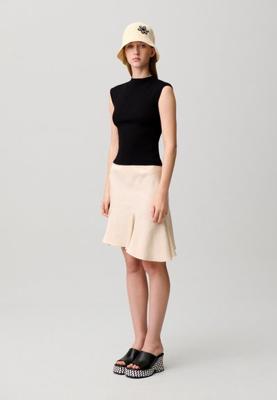 Donna che indossa un top nero senza maniche, gonna svasata color crema, cappello a secchiello crema con motivo nero e sandali neri con plateau e suola decorata.