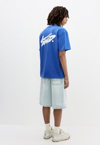 Blå bomulds t-shirt med stort hvidt graffiti-lignende logo på ryggen, parret med lyse denim shorts og hvide sportsko.