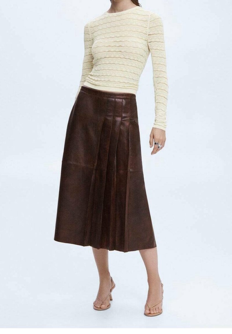 Femme portant un haut texturé crème à manches longues, une jupe midi en cuir plissée marron foncé, et des sandales à talons beige, debout sur un fond blanc.