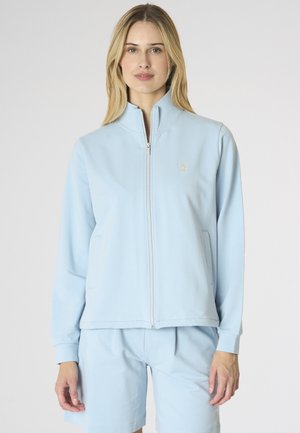 Donna che indossa una giacca azzurro chiaro con zip e pantaloncini abbinati, in piedi davanti a uno sfondo grigio uniforme.