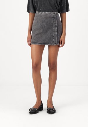 Vrouw draagt een zwarte acid-wash denim minirok, een zwart topje met korte mouwen en zwarte lage schoenen met zilveren gespsluiting.