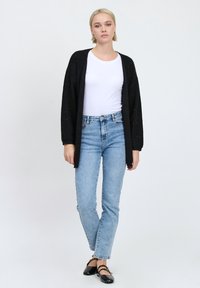 Schwarze Strickjacke, lange Ärmel, offene Vorderseite, über einem weißen T-Shirt getragen. Hellblaue, hoch taillierte Jeans, gerade Passform, zusammen mit schwarzen Ballerinas getragen.