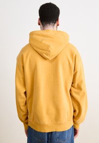 Sweat-shirt à capuche jaune avec une coupe décontractée, des poignets et un ourlet côtelés. Le tissu semble doux, avec une texture lisse.