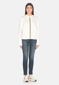 Hoodie blanc avec une fermeture éclair à l'avant et des rayures, associé à un jean bleu ajusté et des baskets blanches. Il a des manches longues et une coupe décontractée.