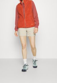 Gilet di pile arancione brillante, con zip frontale, abbinato a una maglietta rossa a maniche lunghe, pantaloni corti beige e scarpe da ginnastica azzurro chiaro.