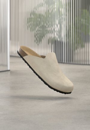Μπεζ παντόφλα τύπου slip-on με μαύρη σόλα, που αιωρείται πάνω από ένα τσιμεντένιο δάπεδο, με θολά φυτά και κάθετες περσίδες στο φόντο.