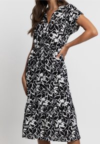 Schwarzes und weißes Blumenkleid mit kurzen Ärmeln, Kragen und Kordelzug in der Taille. Es hat einen gestuften Rock und einen weichen, leichten Stoff.