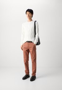 Balta kabelīša adījuma džemperis, sarkanīgi brūni chinos bikses un melni slip-on apavi. Modelis tur melnu plecu somu, stāvot uz baltas fona.