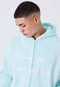 Felpa con zip verde menta, dal tessuto morbido, con un design del logo bianco e cappuccio con cordino, mostrata da un'angolazione laterale.
