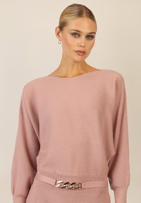 Różowy sweter prążkowany z szerokim dekoltem, oversize'owymi rękawami i dekoracyjnym łańcuszkiem podkreślającym talię. Gładka faktura.