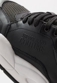 Black Versace Jeans Couture sneaker with textured leather, mesh upper, black laces, and thick white sole, showcasing embossed brand logo.