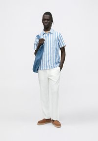 Īsrokas, zilas un baltas svītrainas krekls, bēšas bikses, brūnas uzvelkamas kurpes, turot rokās zilu džinsa jaku uz neitrālas fona.