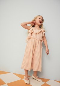 Combinaison rose pâle avec des manches volantées et des jambes plissées, associée à des chaussures plates de couleur claire sur un sol en damier.