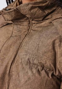 Chaqueta aislante marrón con textura arrugada, cuello alto y detalle de logo en forma de estrella cosido en el pecho. Cierre de cremallera en el cuello.