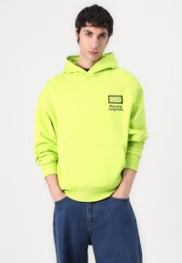 Neongrüner Kapuzenpullover mit vorderer Tasche und schwarzem Logodruck. Gerippte Bündchen und Saum, getragen über blauen Denim-Hosen.