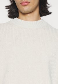 Gros plan d'une personne aux cheveux sombres mi-longs portant un pull en maille côtelée blanche avec un col rond.