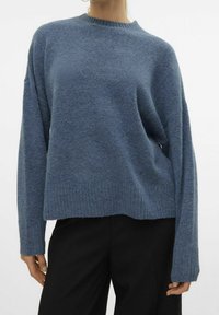 Kvinna som bär en löst sittande, långärmad blå stickad tröja med ribbade muddar och nederkant, ihop med svarta byxor mot en vit bakgrund.