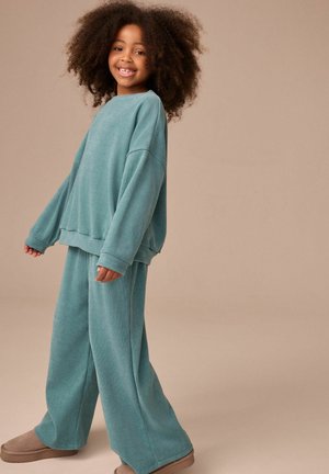 Laps wears a matching teal ribaga pikkade varrukatega särke ja laia jalgadega pükse, koos heledate pehmete sussidega, neutraalse tausta taustal.