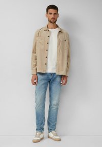 Beige overhemd met knopen en twee borstzakken, gecombineerd met een witte trui en lichtblauwe jeans. Witte sneakers met beige zolen.