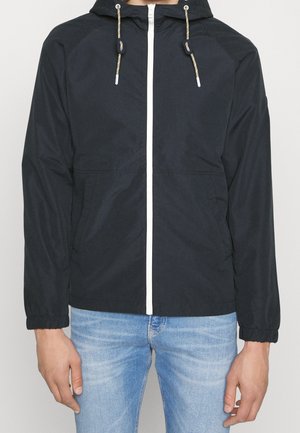 Czarna kurtka z kapturem, z białym suwakiem z przodu i elastycznymi ściągaczami na rękawach, noszona przez osobę w jasnoniebieskich dżinsach, ręce swobodnie opuszczone wzdłuż ciała.