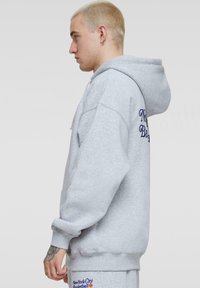 Grå, oversized hoodie i blødt stof med snoretræk i huen. Har broderet tekst på fronten og siden, manchetter på ærmerne og en afslappet pasform.