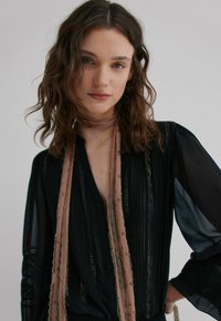 Blouse noire transparente à fines rayures verticales, ornée de dentelle et manches longues ; associée à une écharpe rose décorée de petites perles métalliques.