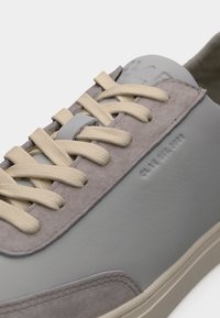 Κοντινό πλάνο ενός γκρι και μπεζ αθλητικού παπουτσιού με σουέντ λεπτομέρειες, μπεζ κορδόνια και ανάγλυφο κείμενο στην πλευρά που γράφει "CLAE EST. 2011".