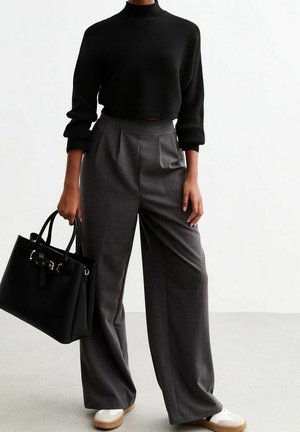 Osoba ubrana w czarny krótki top z długim rękawem, szerokie spodnie z wysokim stanem w kolorze szarym, białe trampki, trzymająca czarną torbę na ramię z detalem klamry.